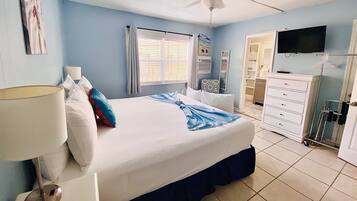 Une chambre à coucher comprenant un lit, un ventilateur de plafond, une fenêtre avec des rideaux, un téléviseur et une commode.