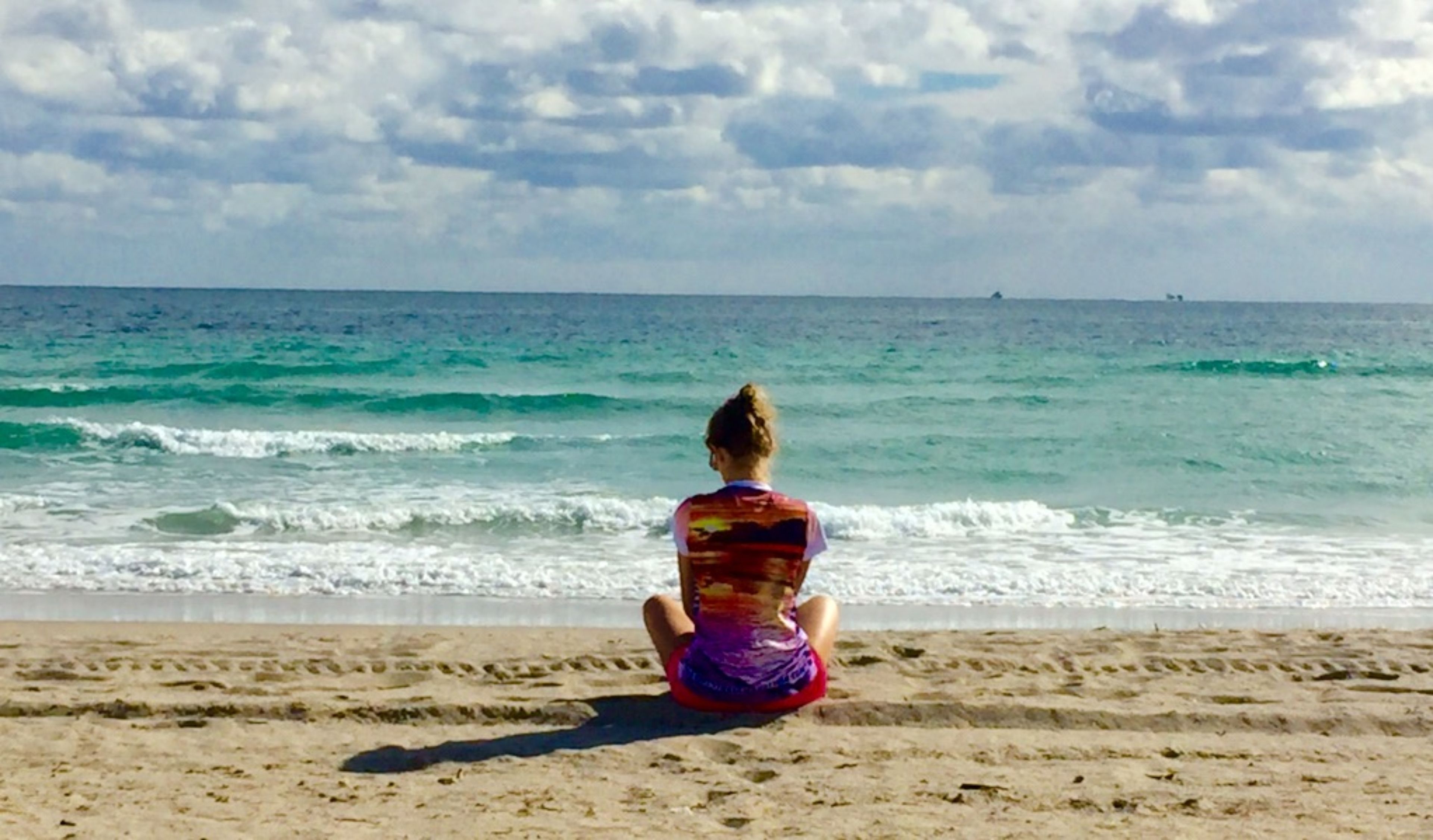 A person sitting on a sandy beach facing the ocean.