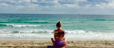 A person sitting on a sandy beach facing the ocean.