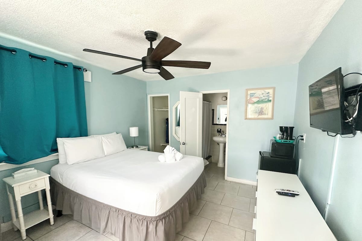 A hotel room with a bed, a desk, a ceiling fan, a TV, and a bathroom visible through an open door.