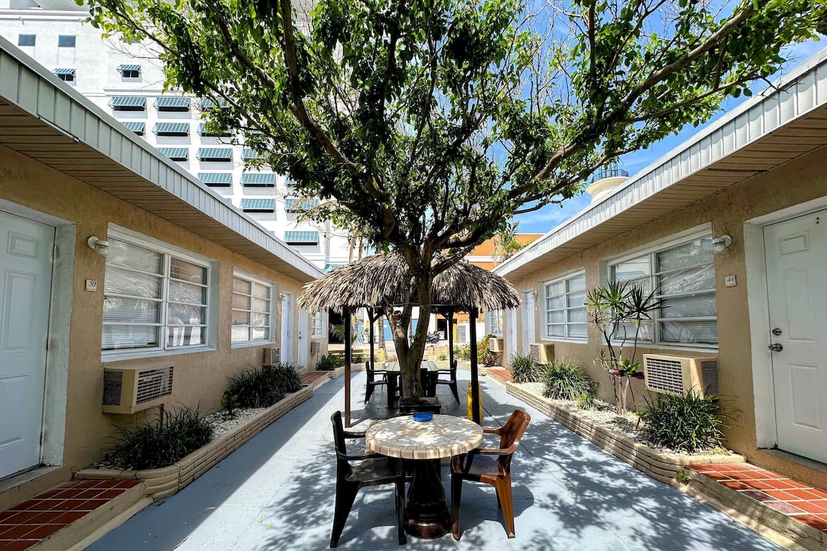 A courtyard with a round table and chairs, surrounded by buildings with numbered doors, a tree providing shade, and a thatched structure.