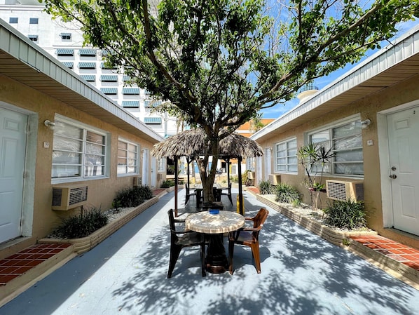 Un patio con una mesa redonda y sillas, rodeado de edificios con puertas numeradas, un árbol que da sombra y una estructura de techo de paja.