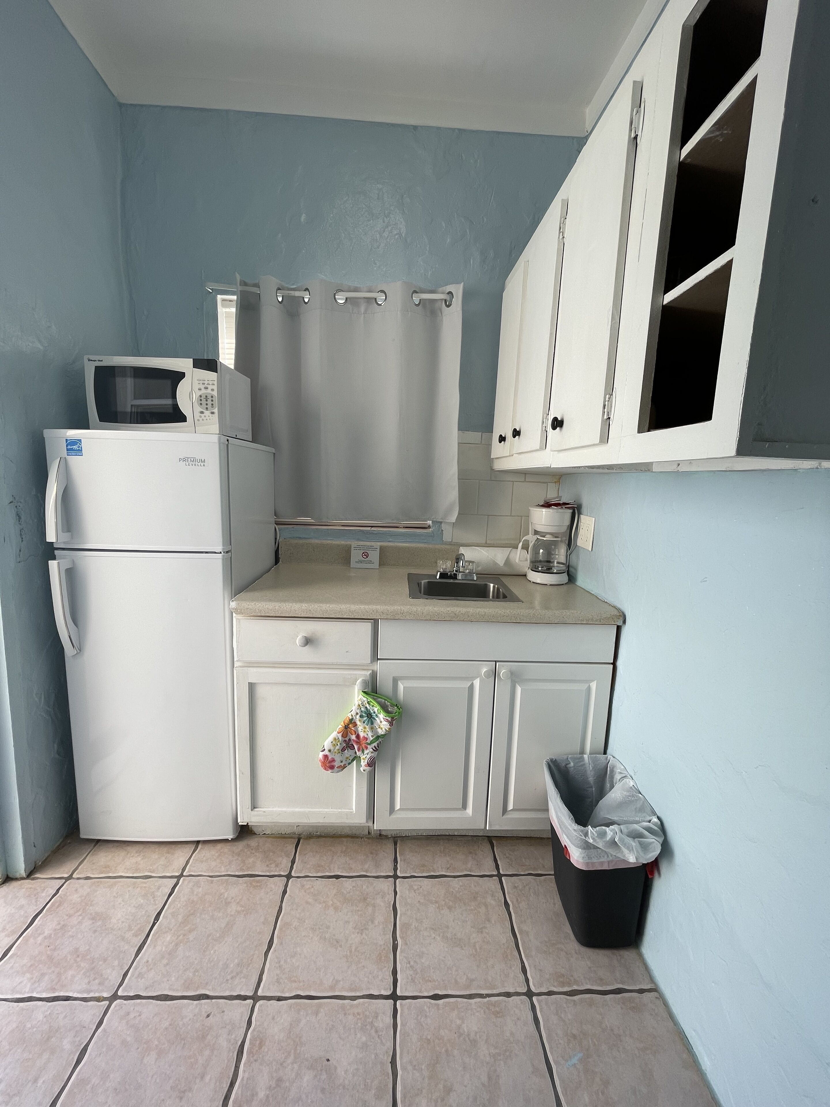 A compact kitchen with white cabinets, a microwave, a refrigerator, a sink, and a trash bin.