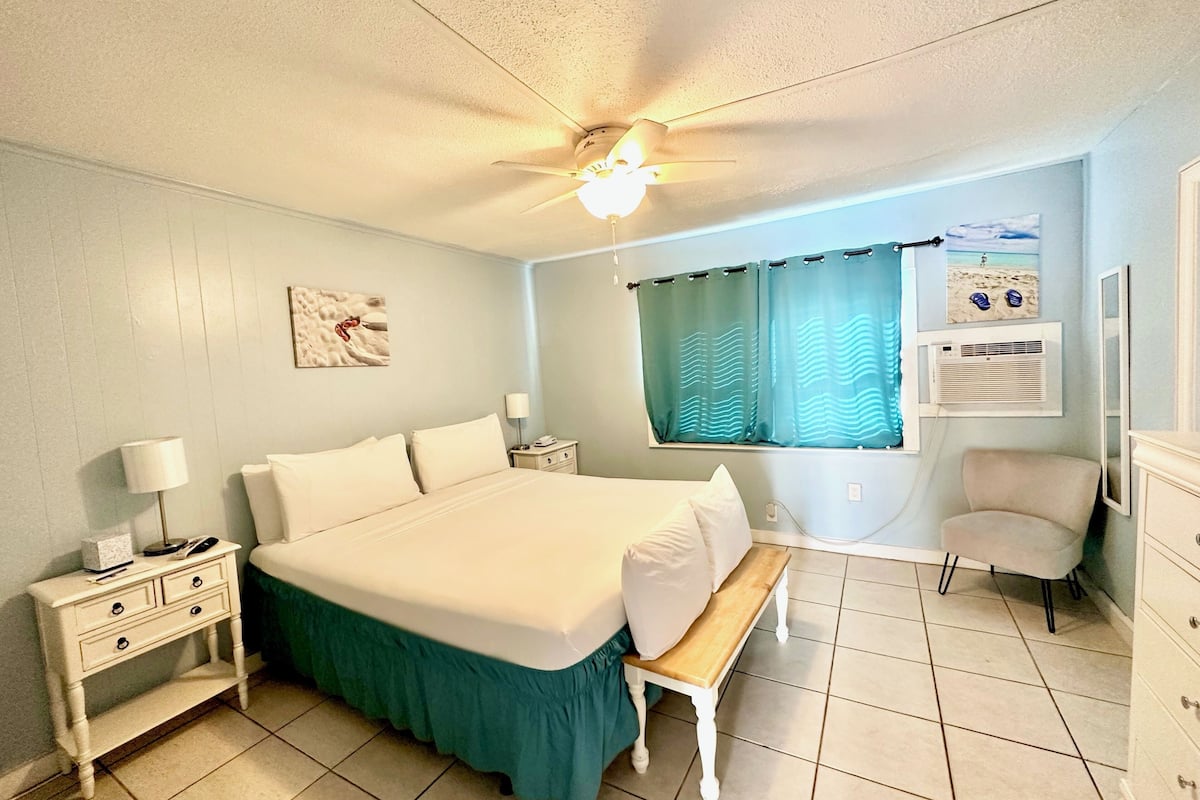 A bedroom with a bed, nightstand, lamp, bench, chair, and a ceiling fan.