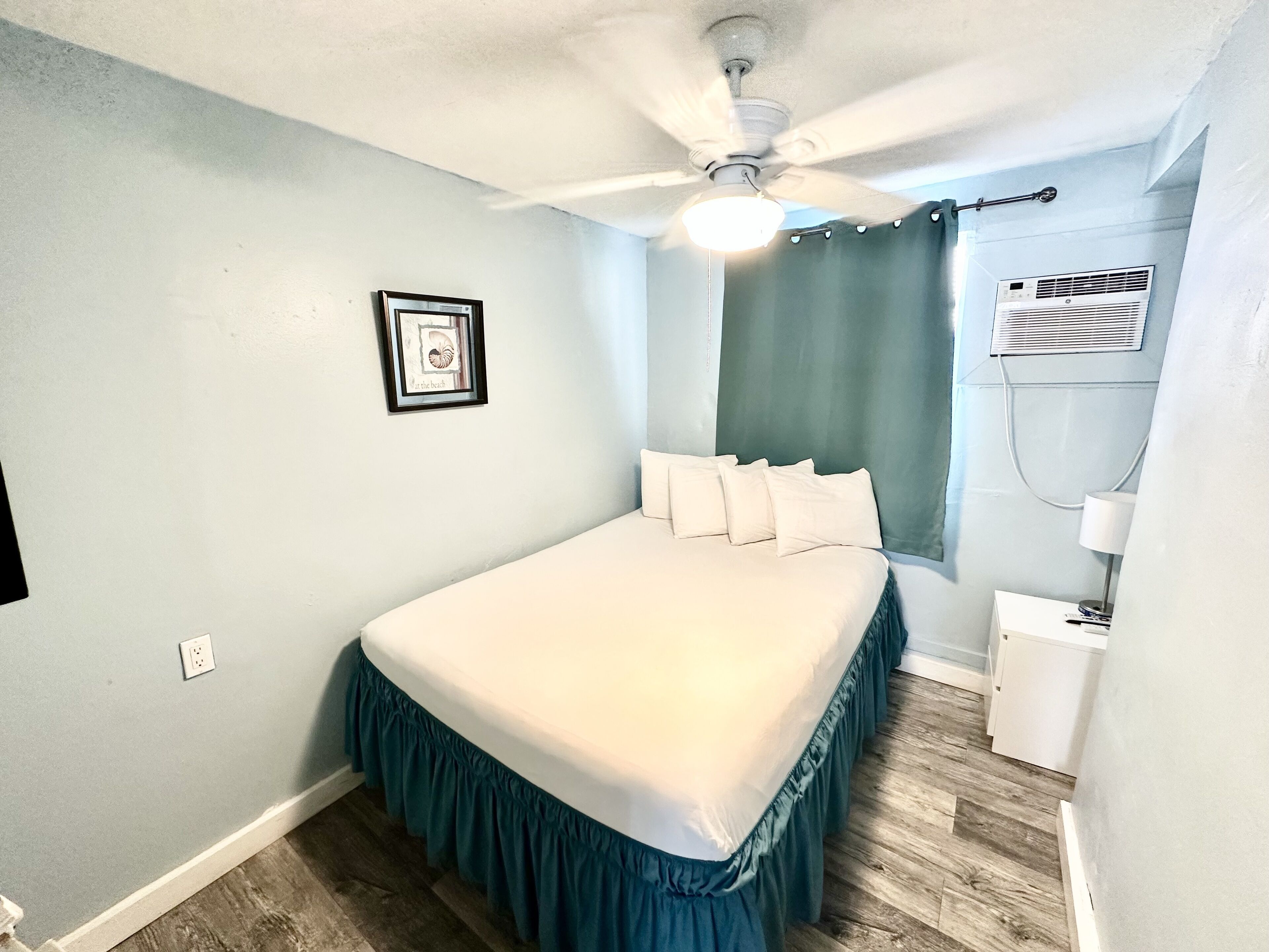 A bedroom with a bed, a ceiling fan, a framed picture, a window with curtains, and an air conditioning unit.