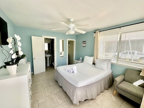 Une chambre avec un lit, un ventilateur de plafond, une fenêtre avec des stores, un petit canapé et une salle de bain visible à travers une porte ouverte.