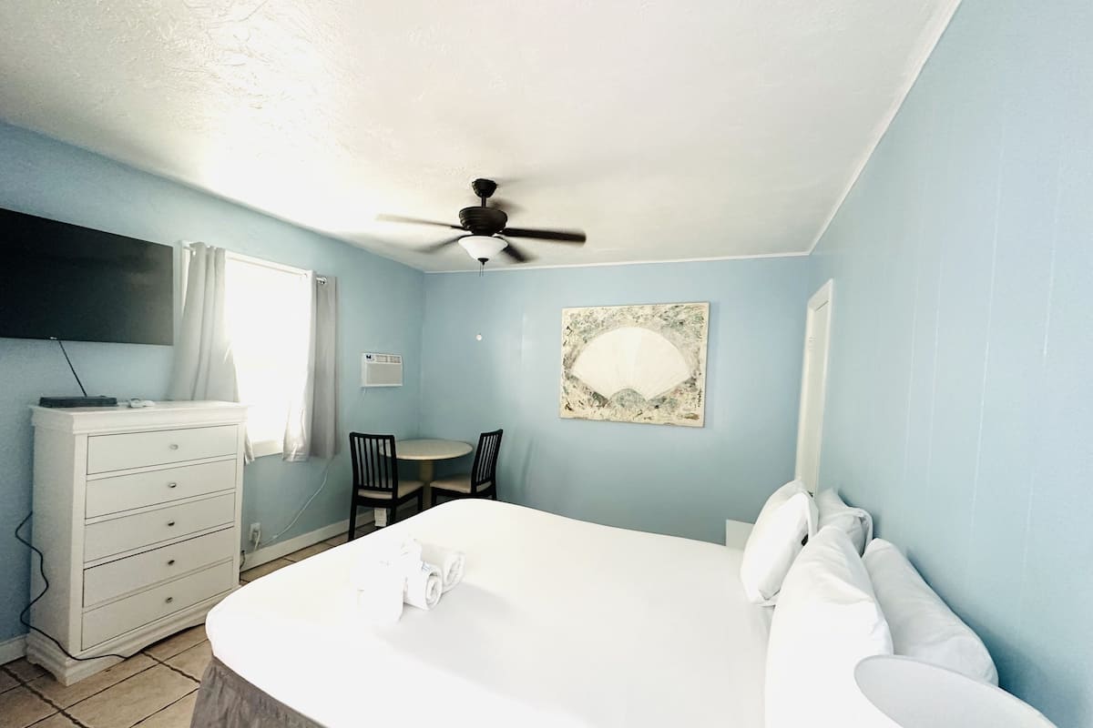 A bedroom with a bed, a desk, two chairs, a ceiling fan, a TV, and a wall-mounted air conditioner.