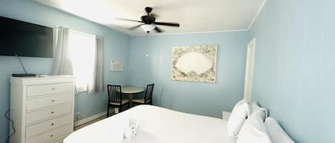 A bedroom with a bed, a ceiling fan, a desk with two chairs, a dresser, and a wall-mounted air conditioner.