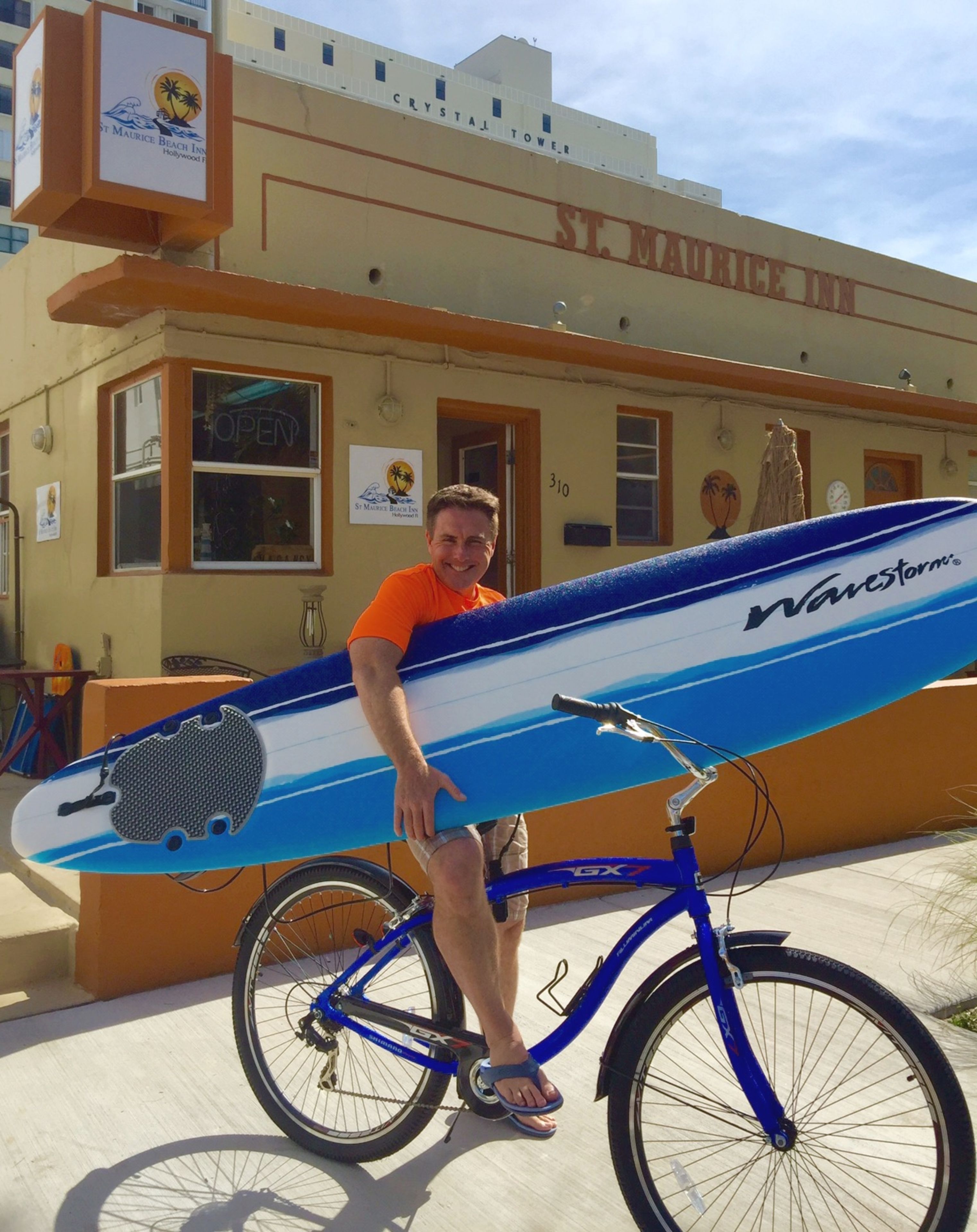 Un hombre de pie junto a una tabla de surf azul y blanca sobre una bicicleta frente a un edificio con la leyenda "St. Maurice Inn".