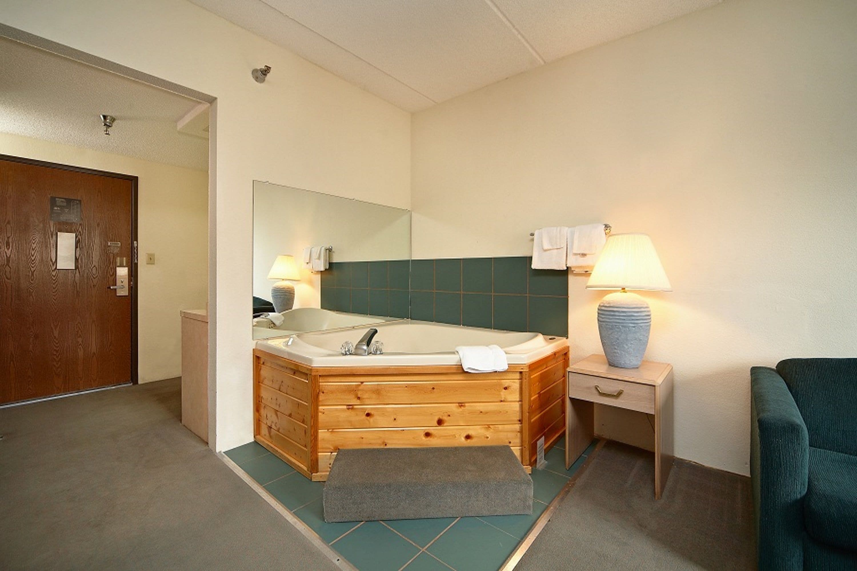A bathroom with a wooden bathtub, a mirror, a towel rack, a small table with a lamp, and a green wall.