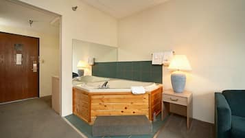A bathroom with a wooden bathtub, a mirror, a towel rack, a small table with a lamp, and a green wall.