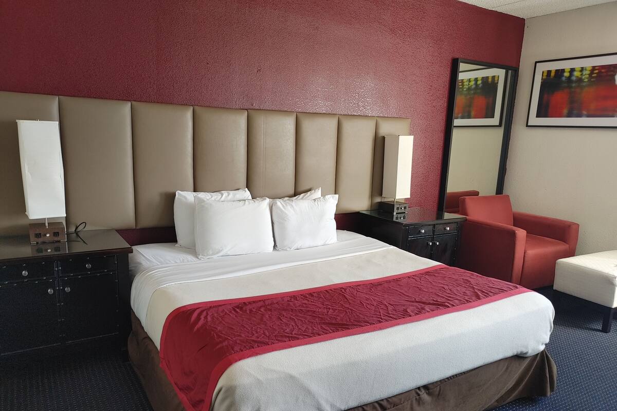 A hotel room with a large bed, a red and white bedspread, a brown skirted bed, a red armchair, a nightstand with a lamp, and a mirror on the wall.