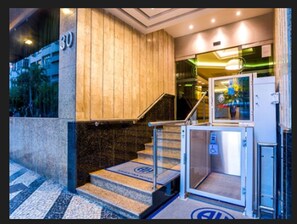 L’entrée d’un hôtel, avec un escalier en marbre, un ascenseur en verre et un panneau indiquant l’entrée.