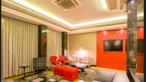 A modern hotel lobby with a glass coffee table, red and grey sofas, a red armchair, and a flat-screen TV on the wall.