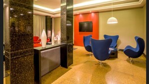 A modern lobby with a reception desk, a flat-screen TV mounted on a red wall, and blue chairs arranged for seating.