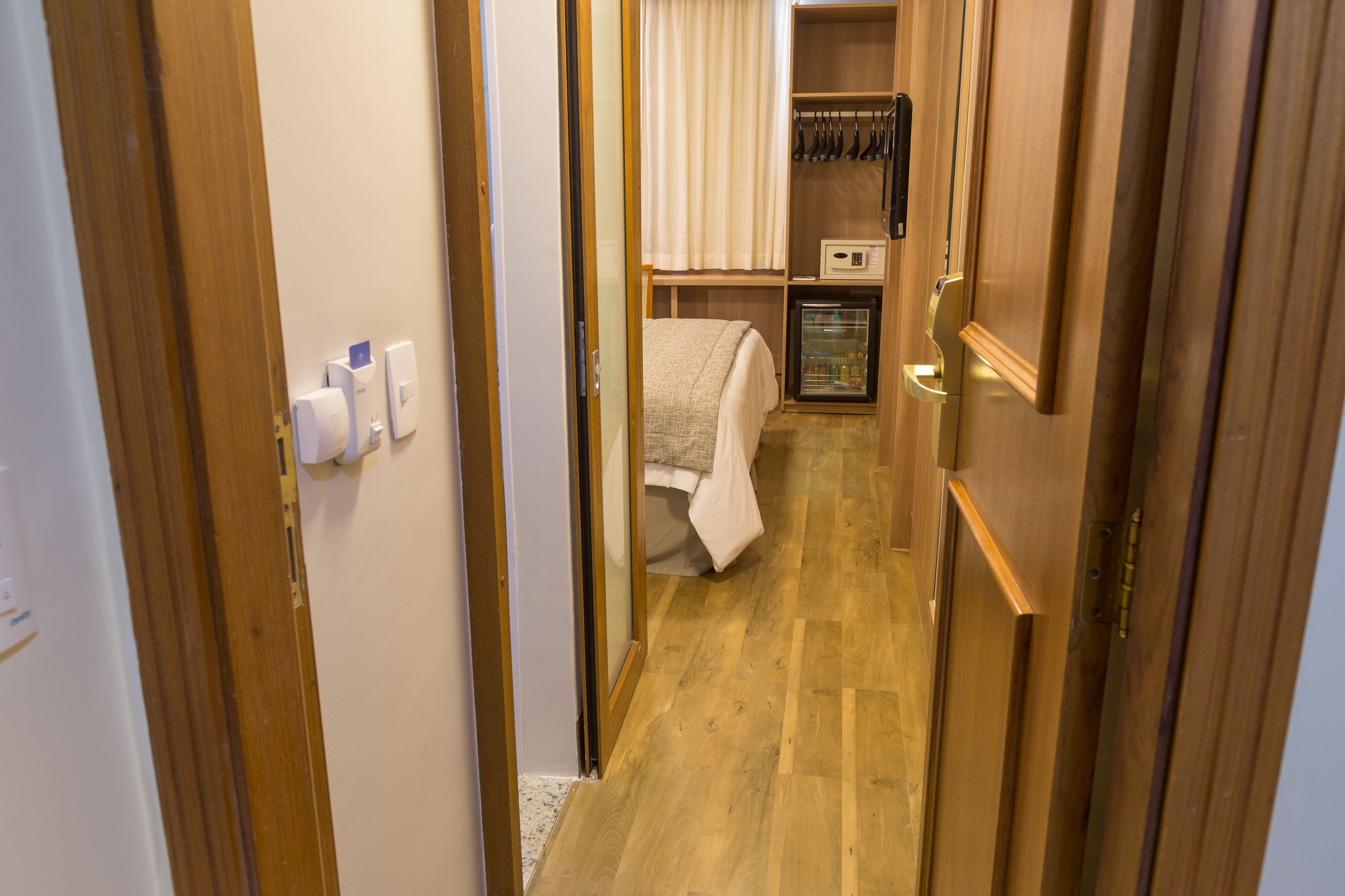 A narrow hallway with wooden flooring, a bed with a white blanket, a small refrigerator, and a shelf with kitchen utensils.