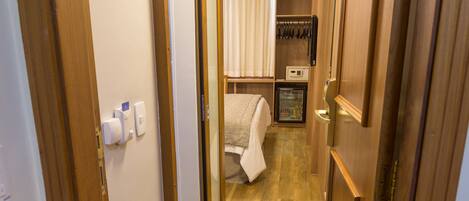 A narrow hallway with wooden flooring, a bed with a white blanket, a small refrigerator, and a shelf with kitchen utensils.