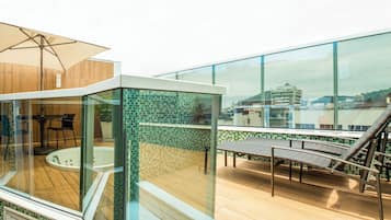 A modern rooftop terrace with a glass-enclosed jacuzzi, a seating area with chairs, and a view of the cityscape.