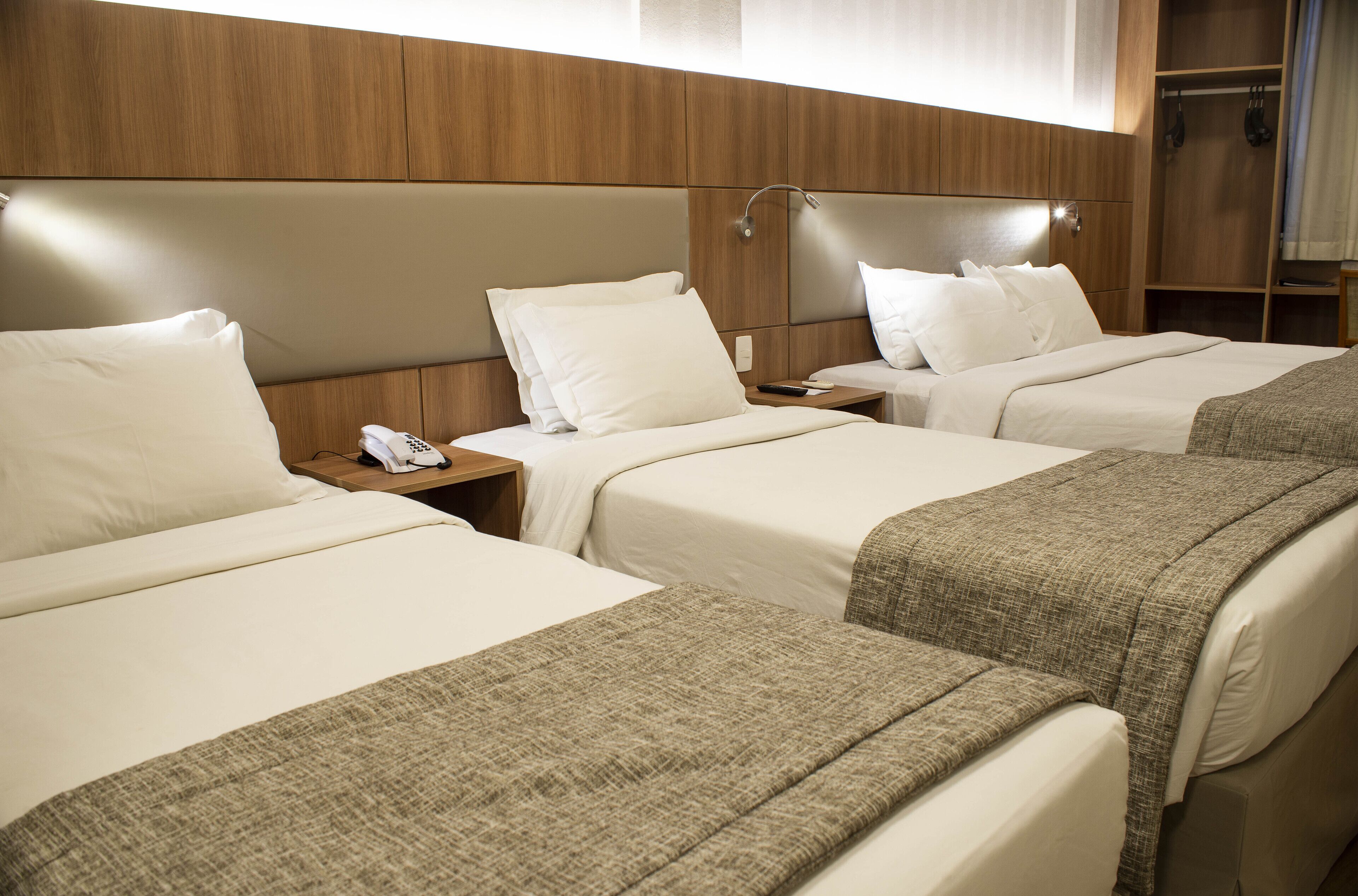A hotel room with three beds, each with white linens and a brown throw blanket. There is a wooden headboard, a bedside table with a phone, and a nightstand with a lamp.