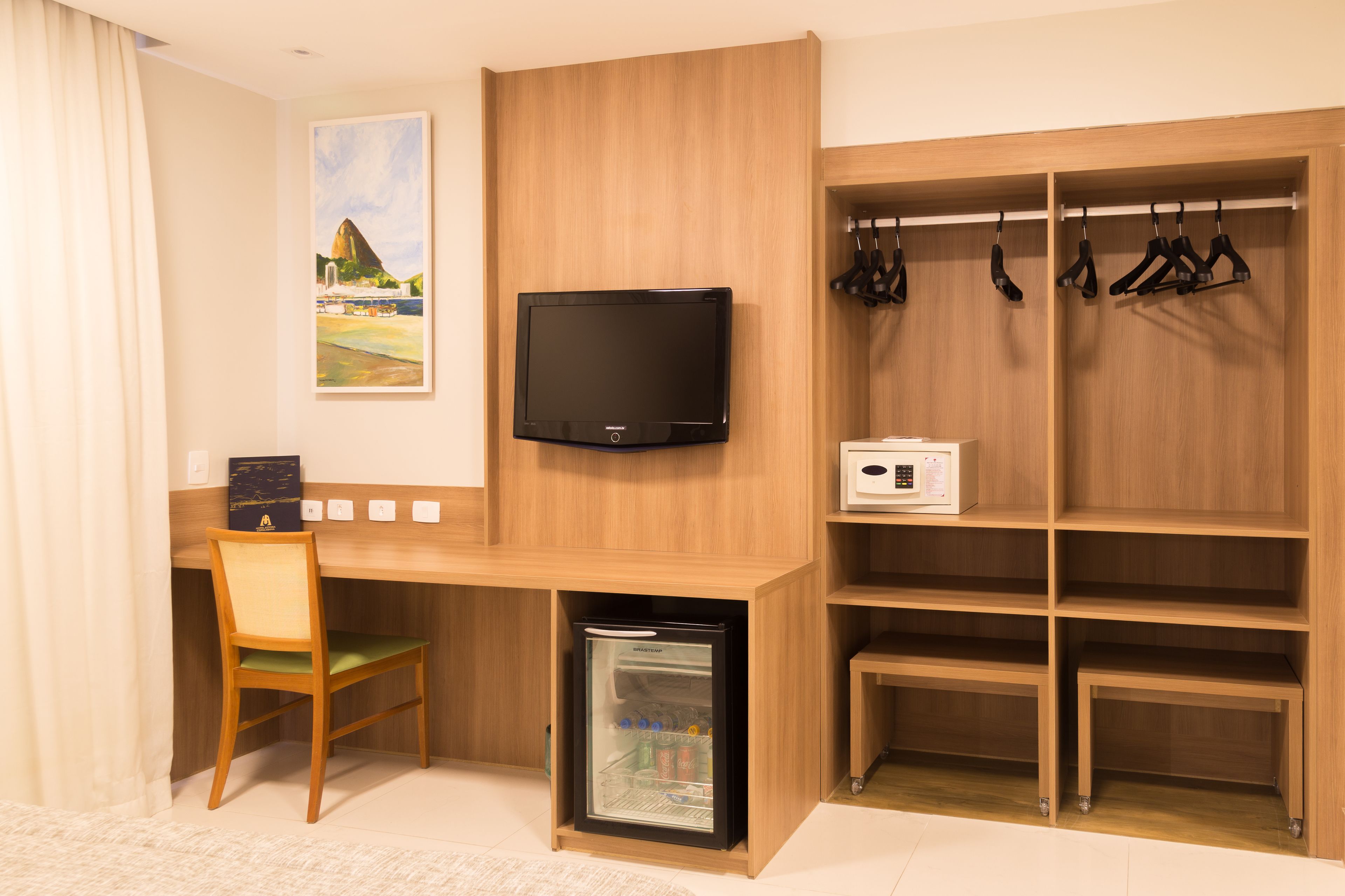 Ein gemütlicher Raum mit Schreibtisch aus Holz, Fernseher, einer kleinen Kühlbox, einem Sessel und einem Kleiderhaken.