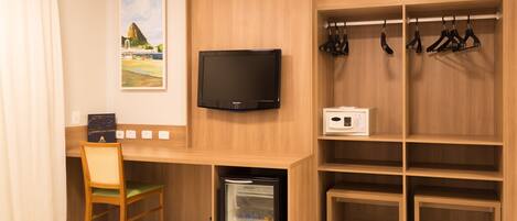A compact room with a wooden desk, a television, a mini-fridge, a chair, and a coat rack.