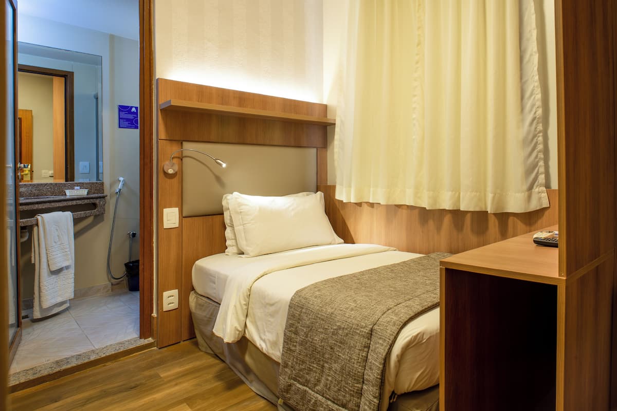 Habitación de hotel con cama, cabecera de madera, mesita de noche, zona de ducha y baño con espejo y perchero.