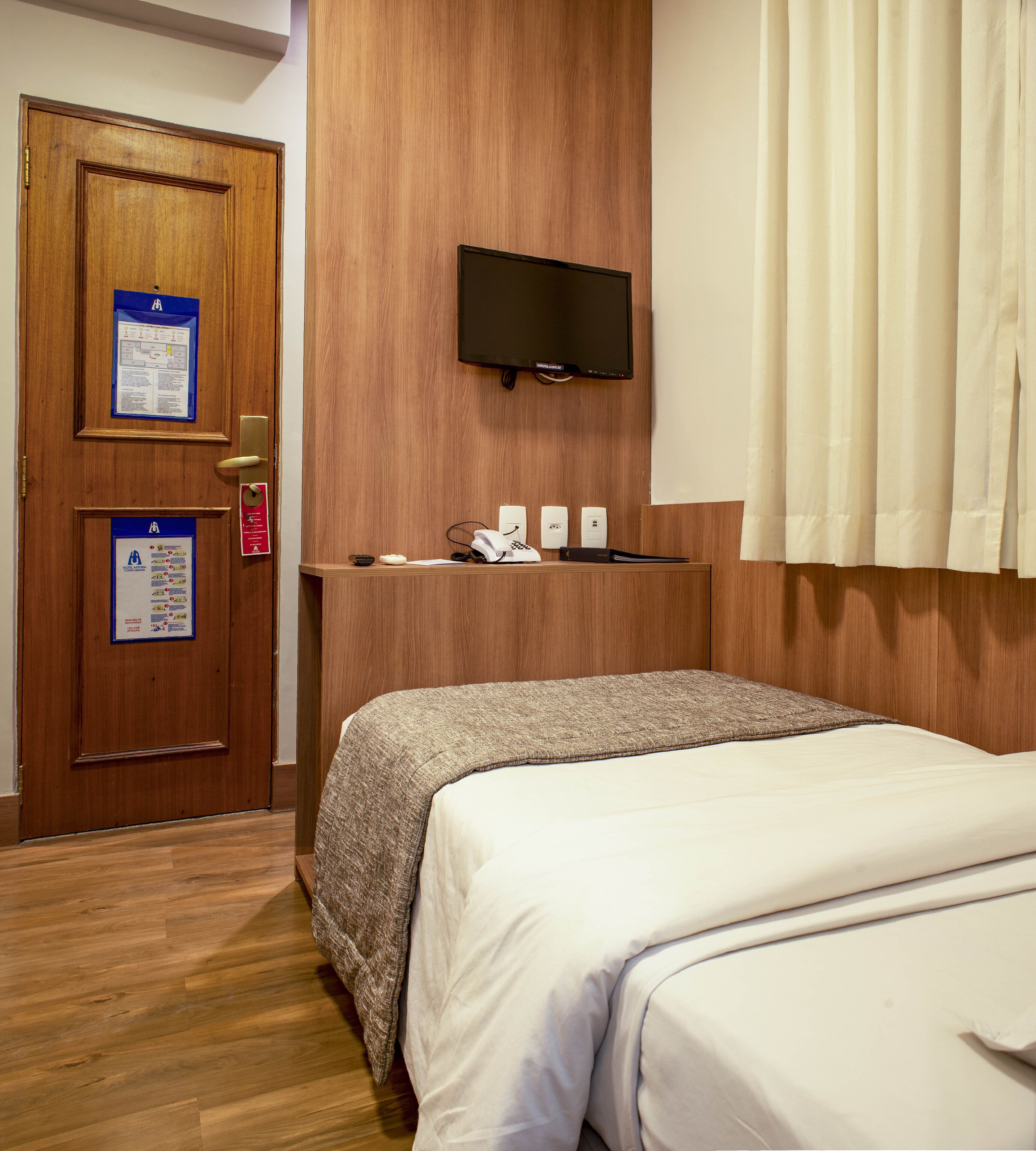 A hotel room with a wooden door, a mounted flat-screen TV, a desk with a telephone, and a bed with a textured blanket.