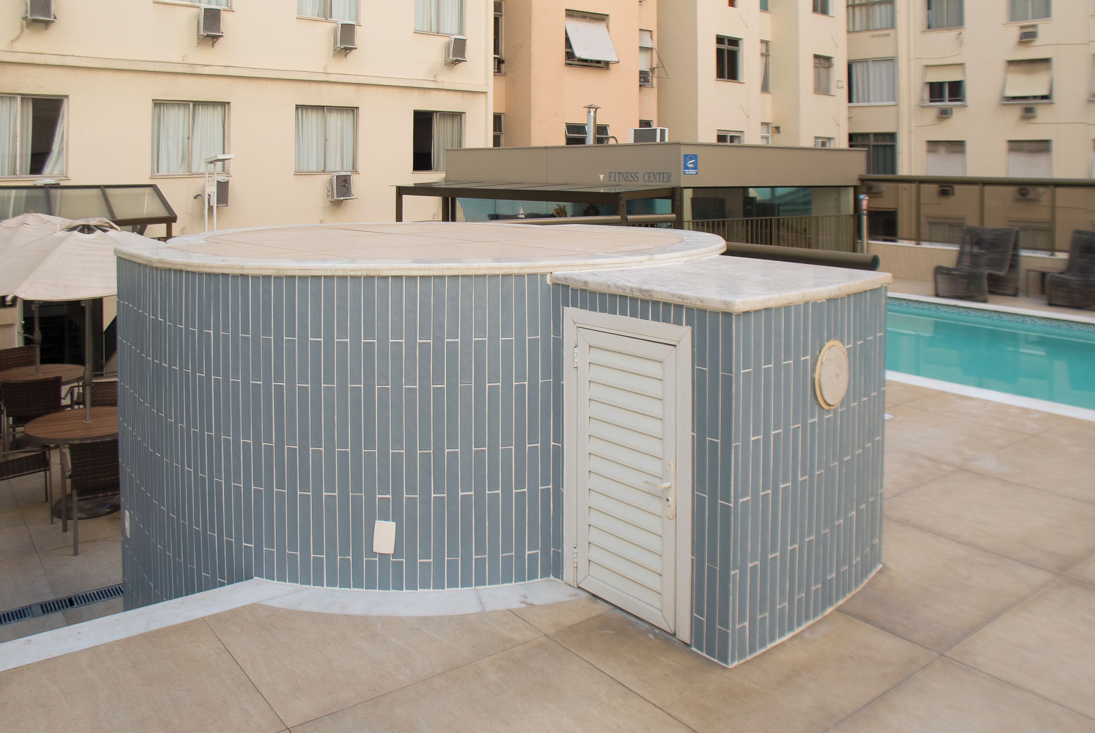 Uma área na piscina com uma banheira de hidromassagem azulejada, assentos externos e um centro de fitness ao fundo.