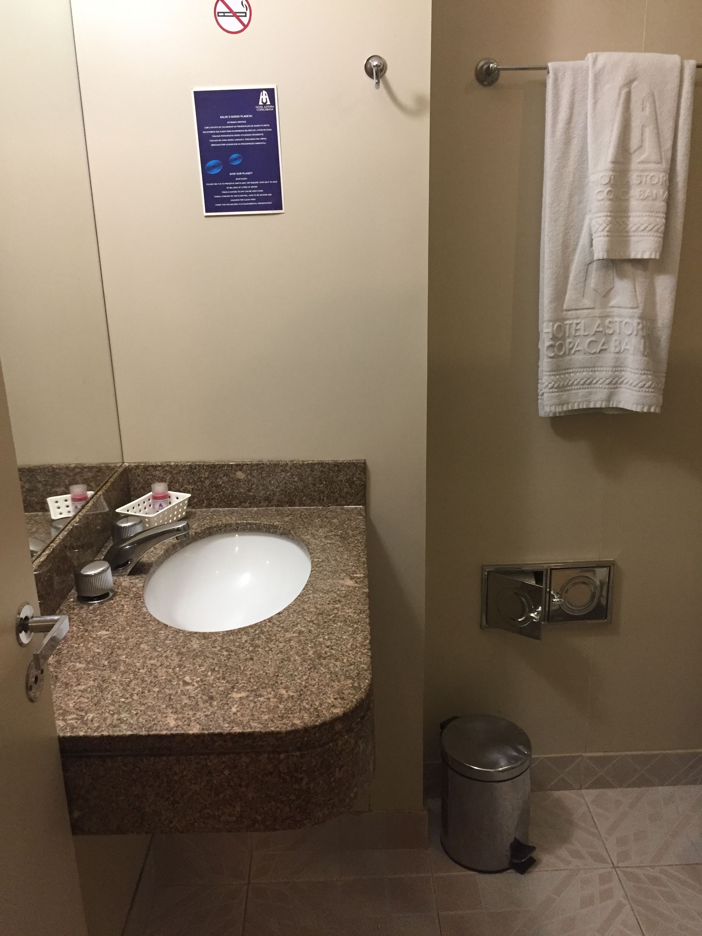 Un baño con lavamanos de granito, un toallero con una toalla blanca, un cubo de basura y un cartel de prohibido fumar.
