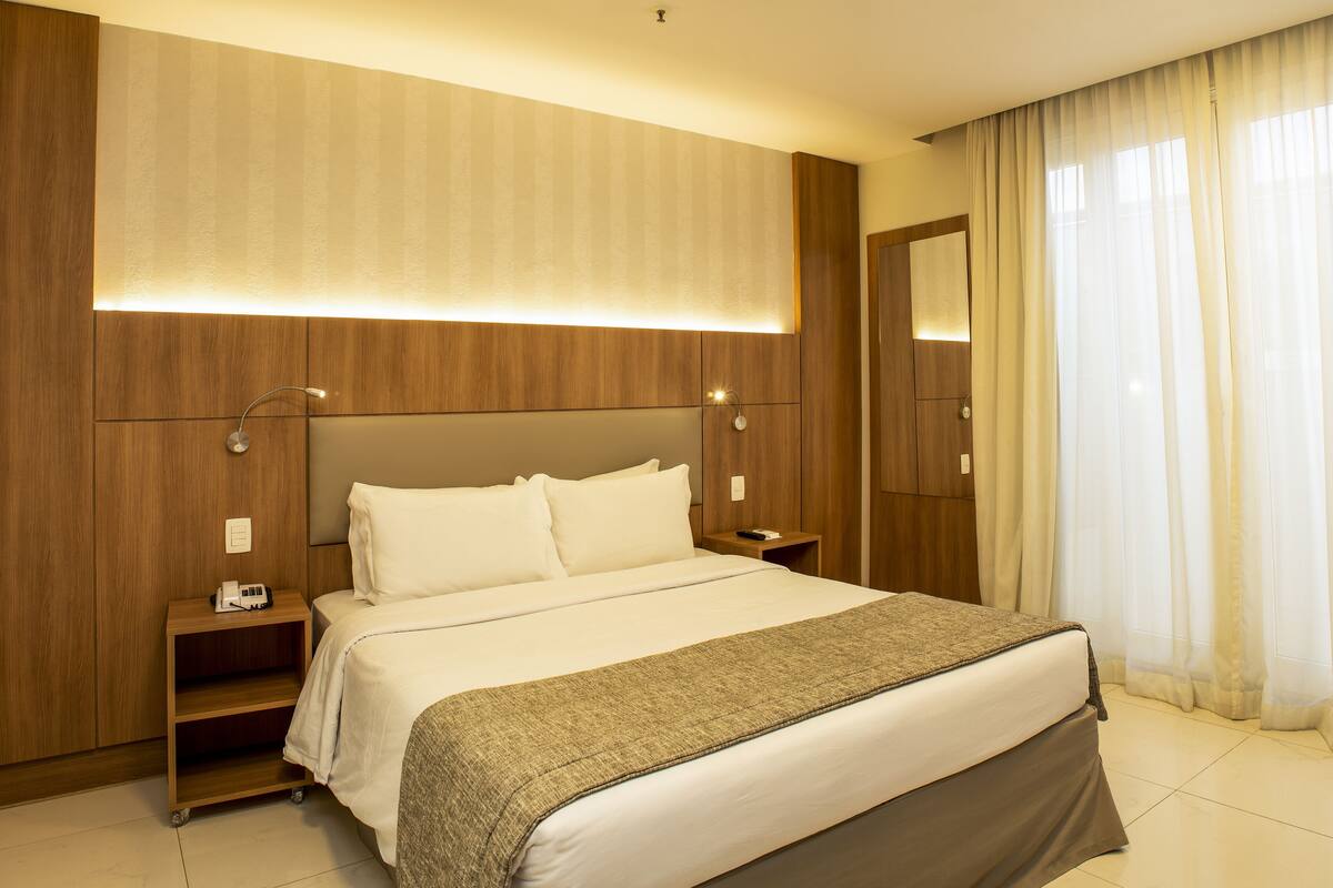 Una habitación de hotel con una cama grande, cabecera de madera, mesitas de noche con lámparas, un estante pequeño y un espejo amplio.