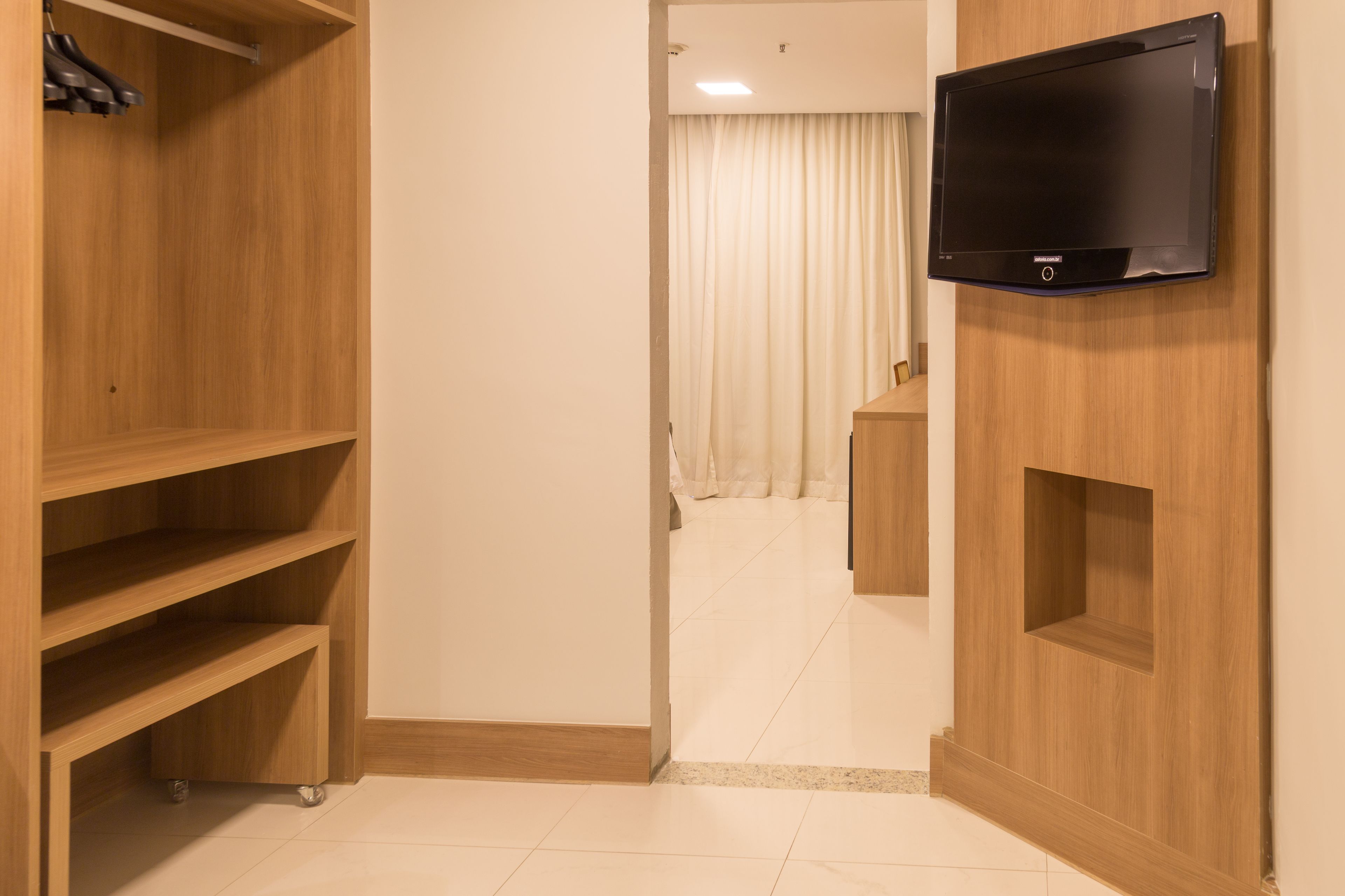 Habitación de hotel con un armario de madera, un televisor de pantalla plana montado en la pared y un pasillo que lleva a un baño con cortina.