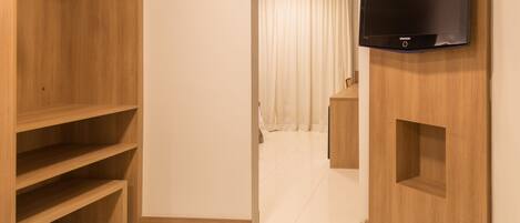 Habitación de hotel con un armario de madera, un televisor de pantalla plana montado en la pared y un pasillo que lleva a un baño con cortina.