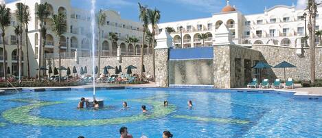 A pool area with multiple people swimming and relaxing, surrounded by a large resort building with multiple balconies and palm trees.