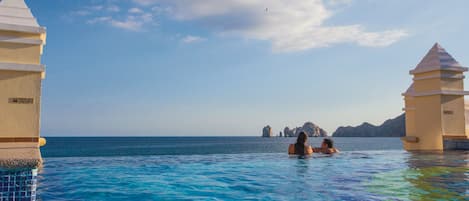 Una piscina azul cristalina con dos personas nadando, rodeada de pilares blancos y vistas al mar y a islas distantes.