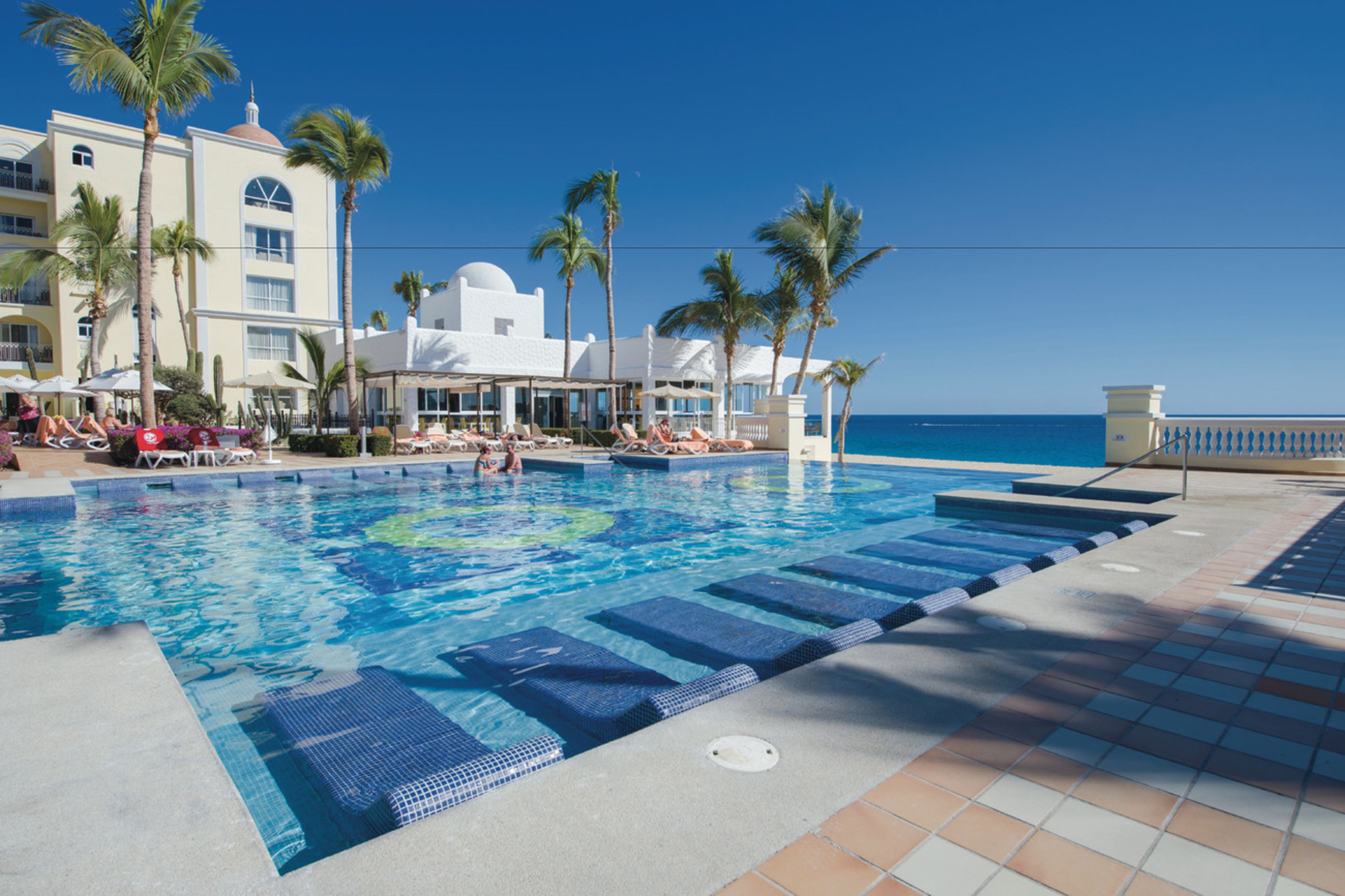 A pool area with lounge chairs, palm trees, and a view of the ocean.