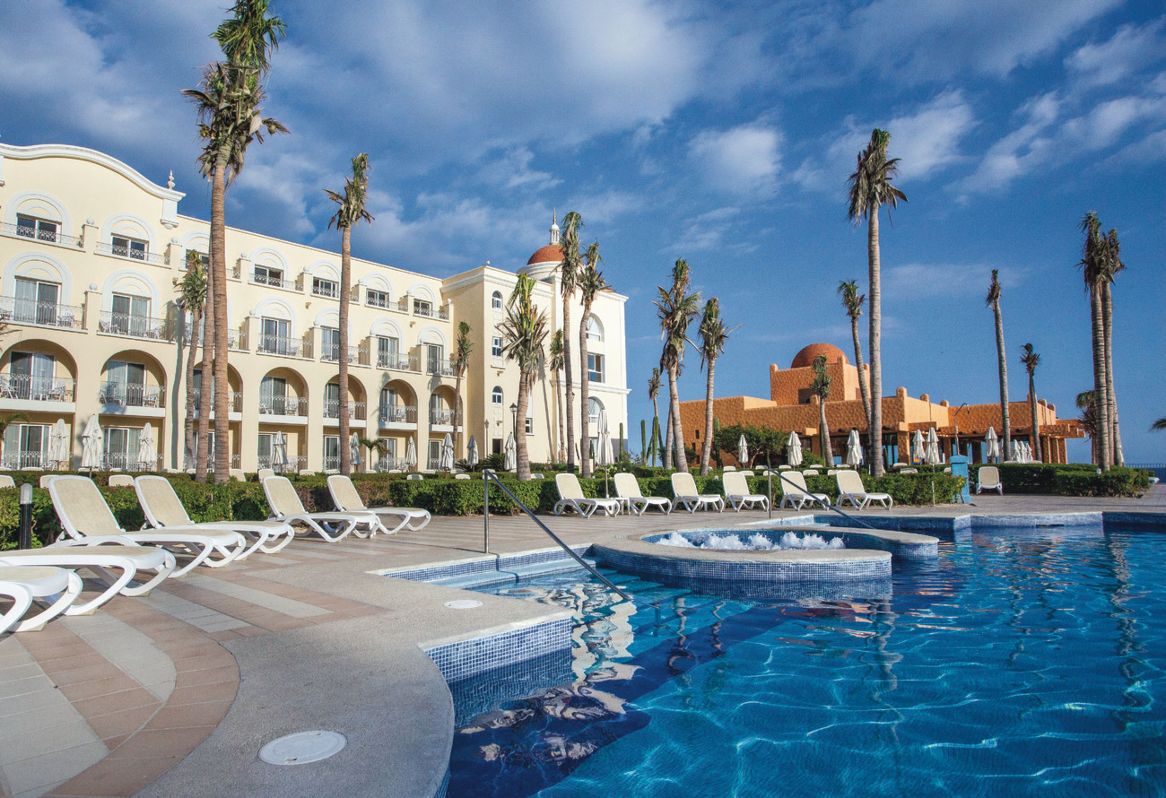 Zona de piscina de hotel con sillas de descanso, palmeras y un edificio grande con balcones.
