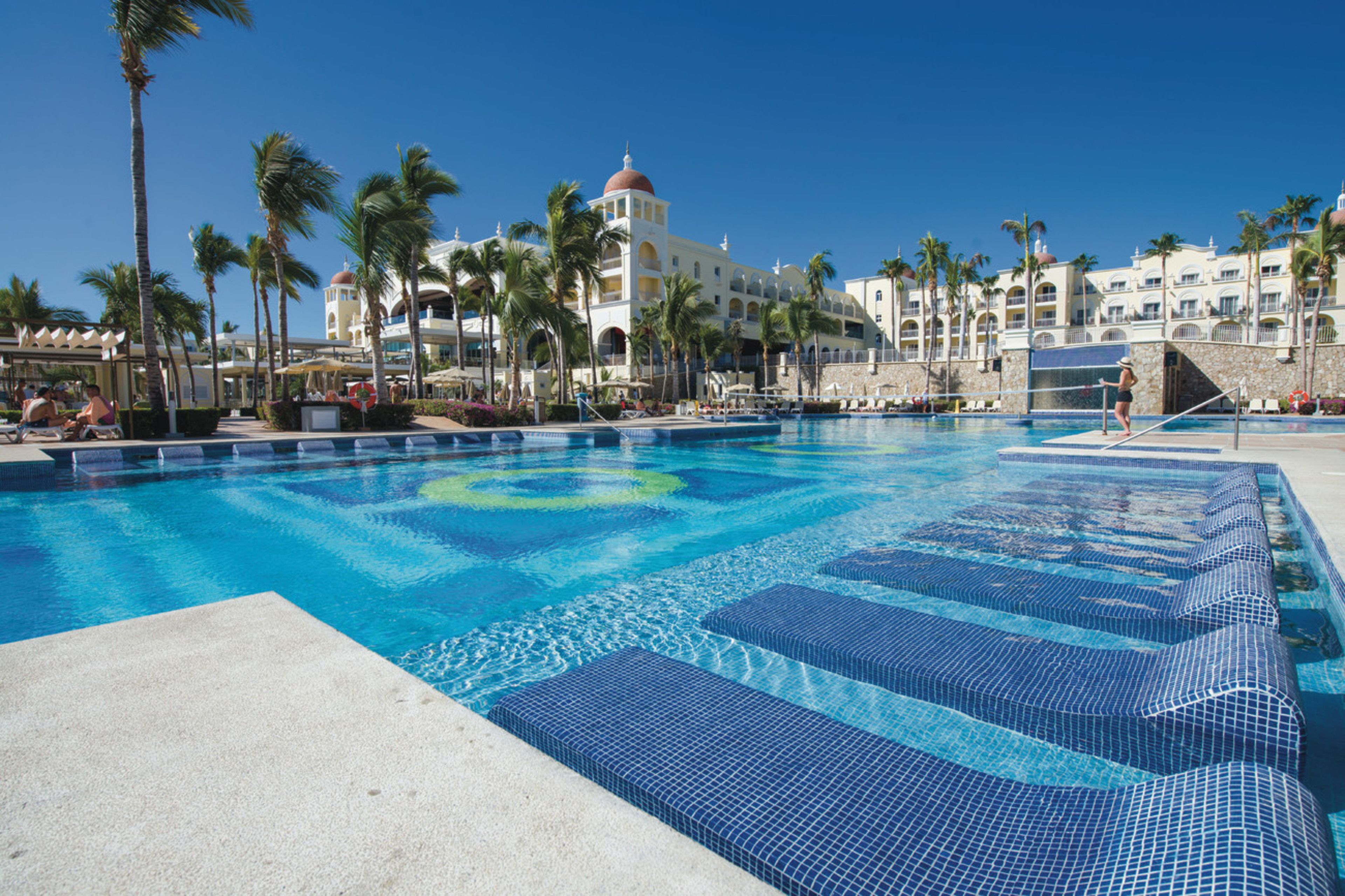 Una gran piscina con mosaicos azules, rodeada de palmeras y un hotel con varios balcones y un edificio de forma abovedada.
