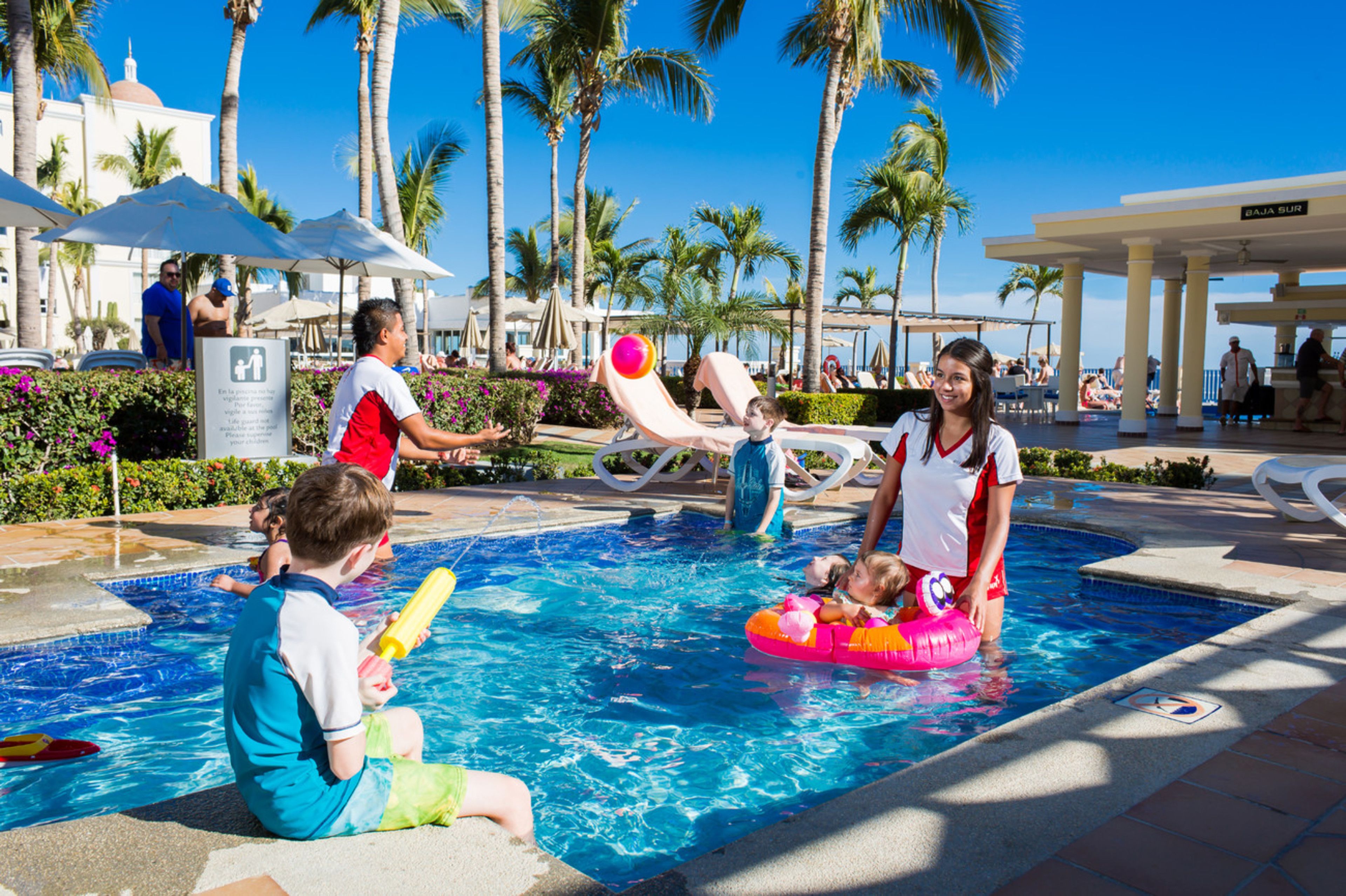 A swimming pool area with palm trees, umbrellas, and people.