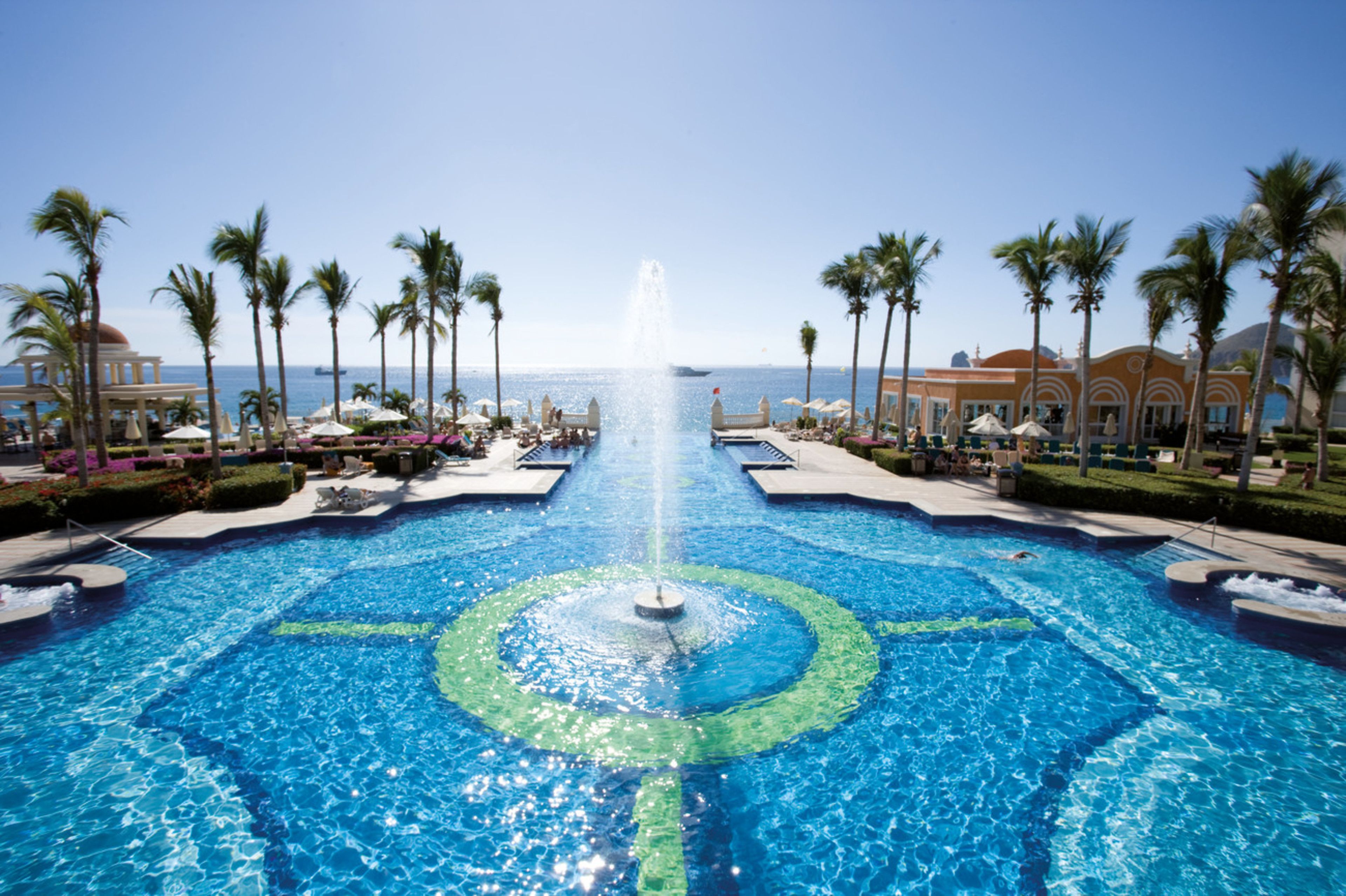 A large pool with a central fountain, surrounded by palm trees and lounge chairs, with a view of the ocean in the background.