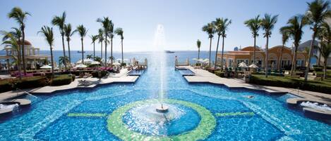 A large pool with a central fountain, surrounded by palm trees and lounge chairs, with a view of the ocean in the background.