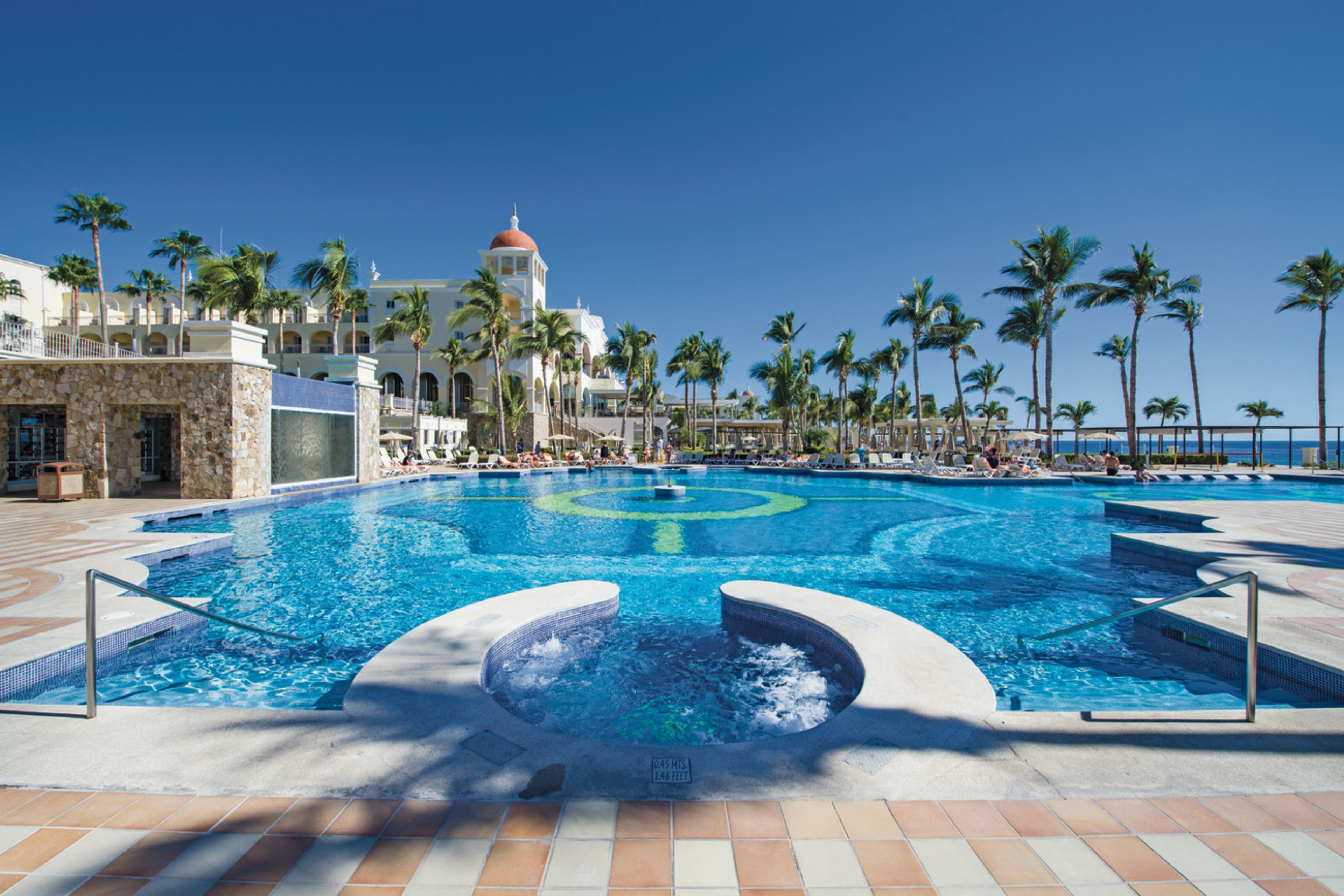 A large outdoor swimming pool with a hot tub in the center, surrounded by palm trees and a building with a red dome.