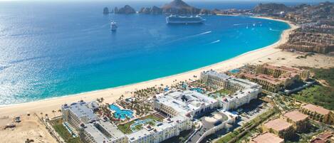 Vista aérea de un hotel costero con varias construcciones, una playa y un gran crucero al fondo.