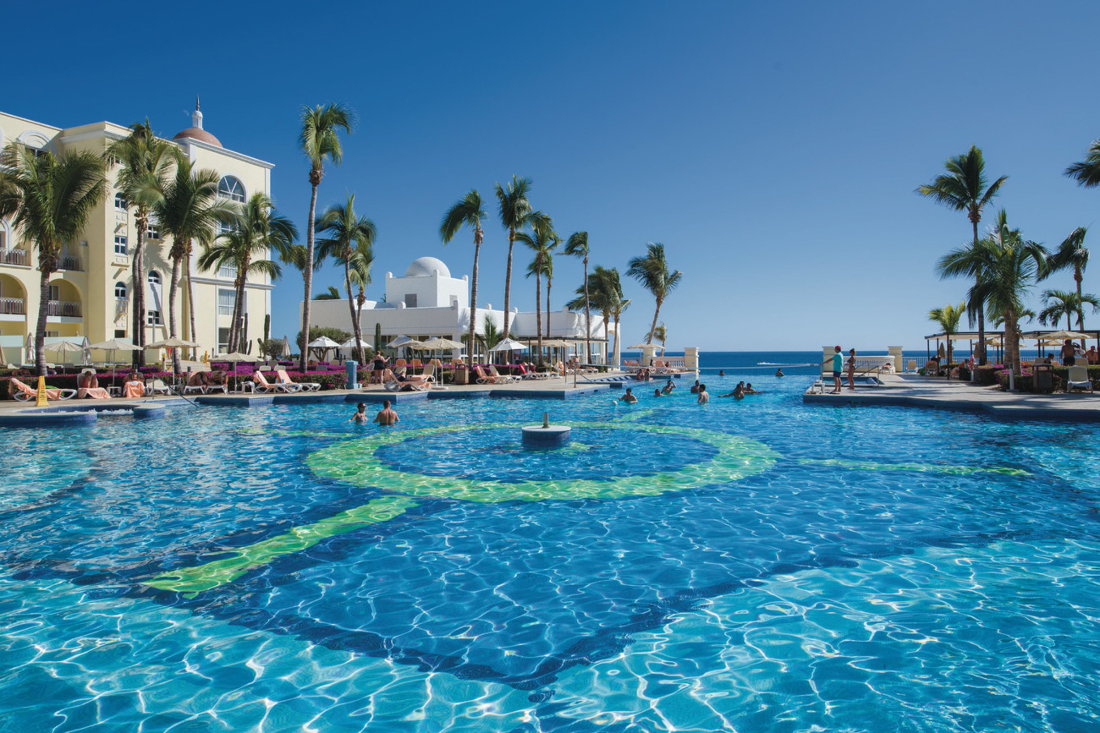 A large outdoor swimming pool surrounded by palm trees and a hotel building with multiple balconies.
