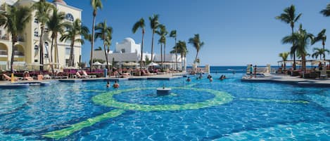 En stor udendørs swimmingpool omgivet af palmer og en hotelbygning med flere balkoner.