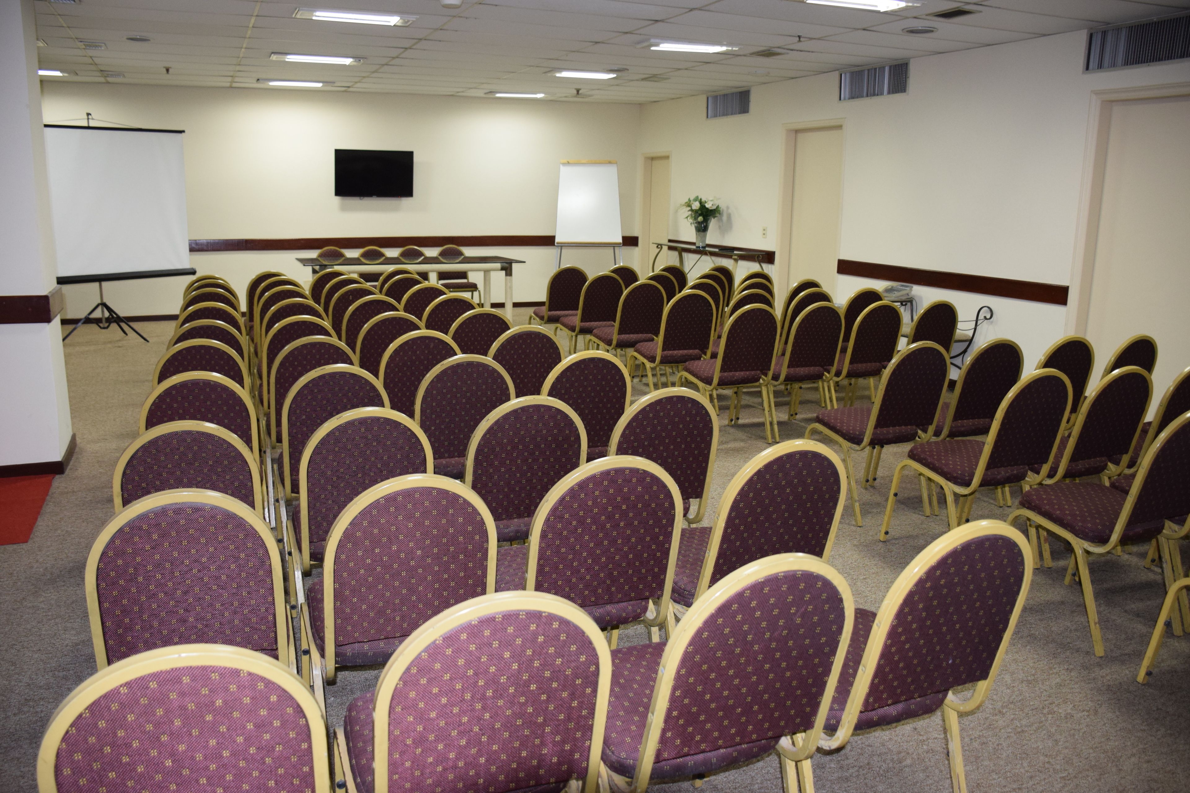 Un salón de conferencias con hileras de sillas moradas, una pizarra, una pantalla de proyector y un televisor en la pared.