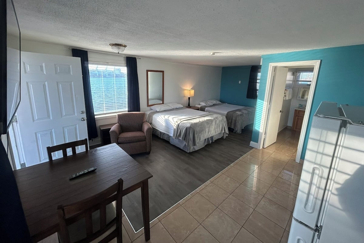 A compact room with two beds, a small table, a chair, a desk, a lamp, and a refrigerator.