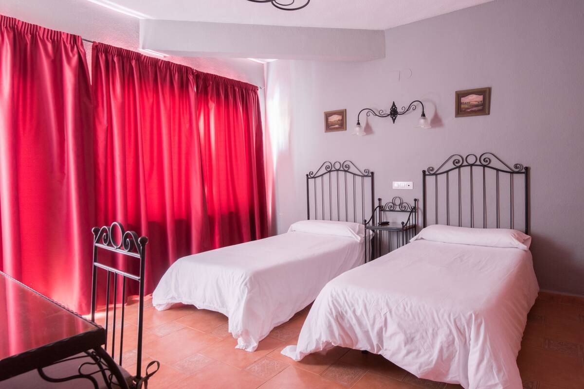 A room with two single beds, a metal chair, a table, red curtains, and a wall-mounted light fixture.