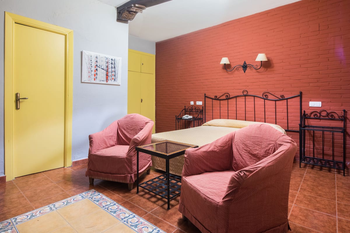 A room with a bed, two pink chairs, a small table, a yellow door, and a red brick wall.