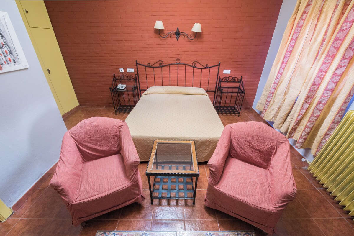 A room with a bed, two chairs, a small table, and a wall-mounted lamp.