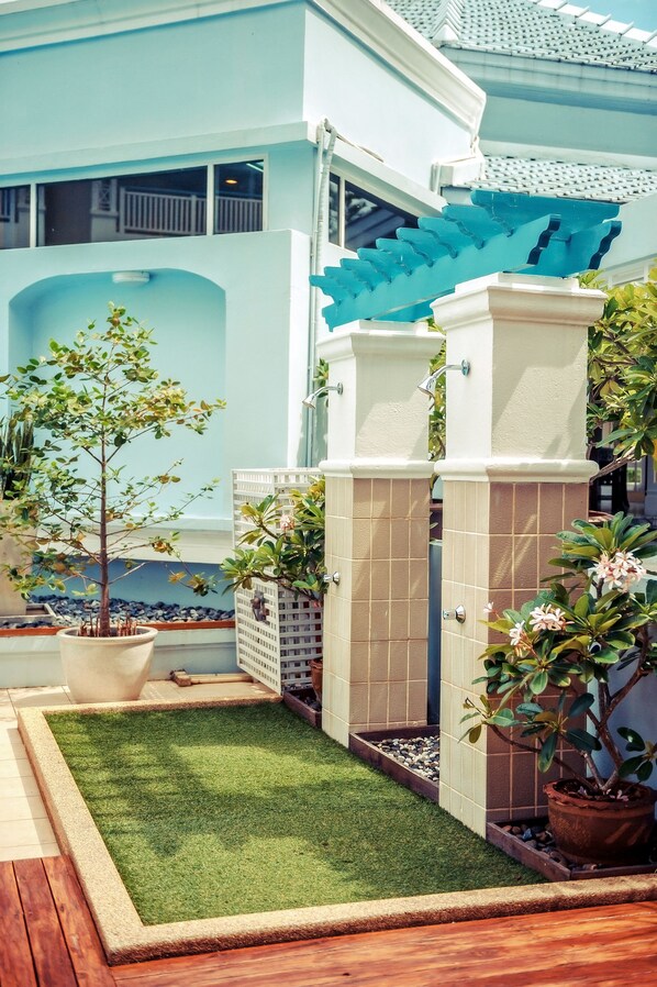 Un edificio moderno con fachada azul claro, un pequeño jardín con plantas en macetas y un camino empedrado que conduce a una entrada con puerta.